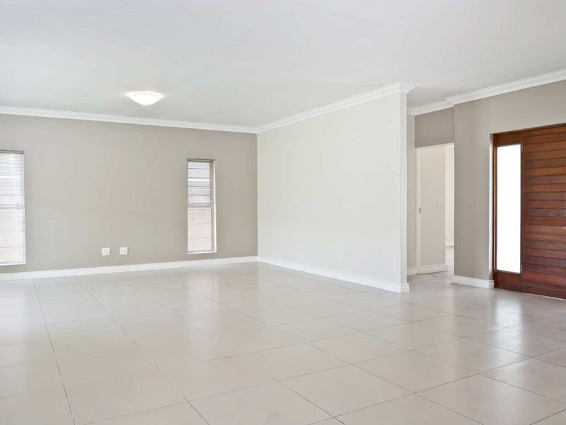 A spacious, bright, and empty living area featuring neutral wall tones, large floor tiles, crown molding, and a solid wooden entrance door.