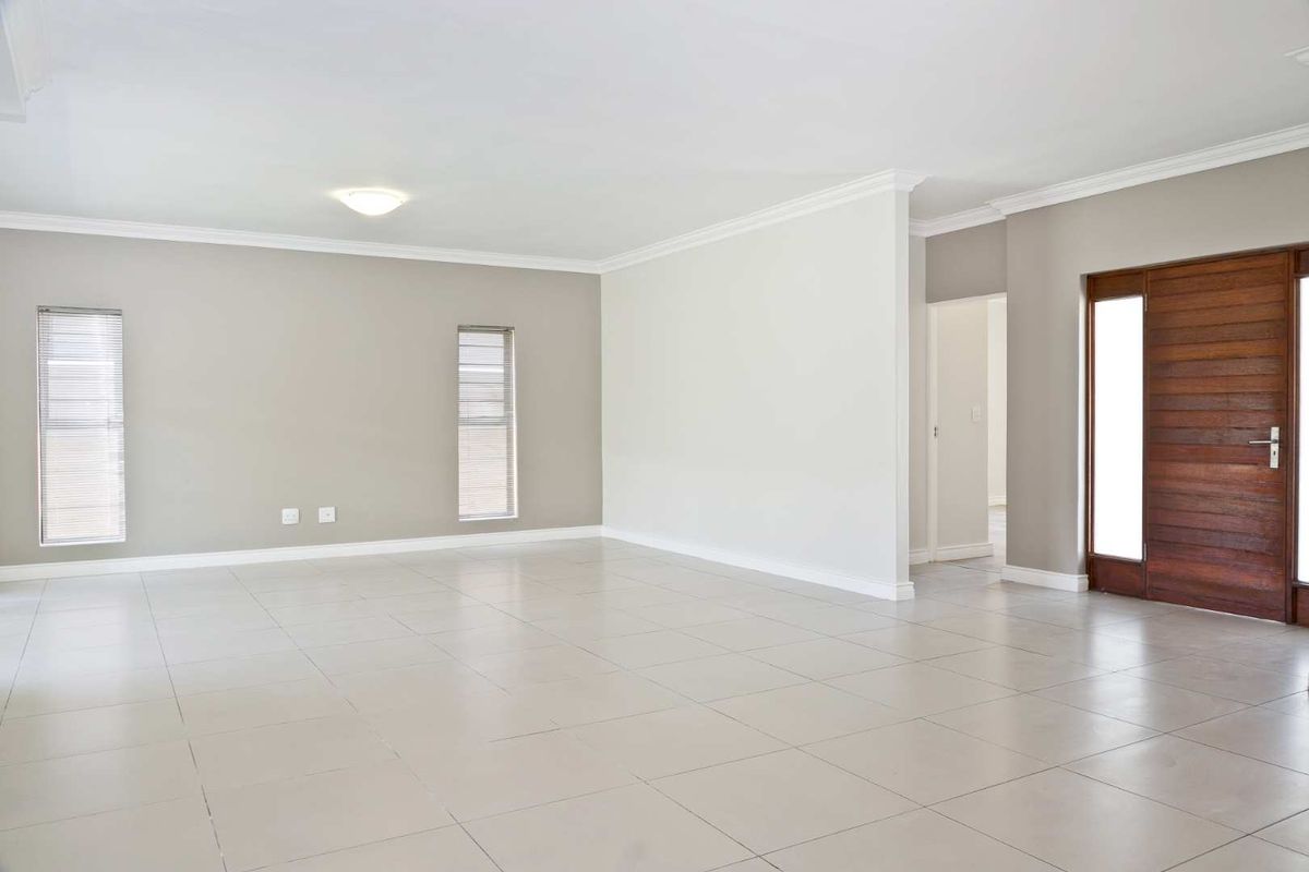A spacious, bright, and empty living area featuring neutral wall tones, large floor tiles, crown molding, and a solid wooden entrance door.