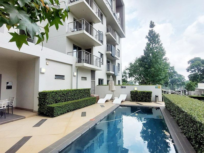 A well-maintained communal swimming pool area featuring a tiled deck, manicured hedges, and views of the modern apartment building facade.