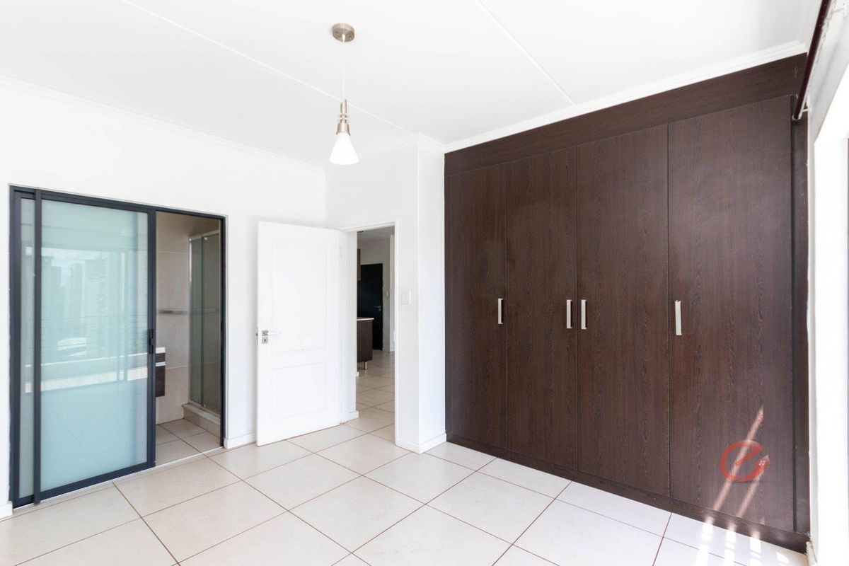 A bright, empty bedroom featuring dark wood built-in cupboards, neutral tiled flooring, and direct access to an en-suite bathroom.