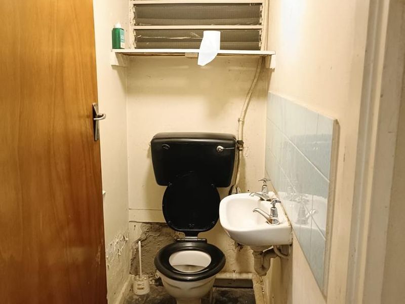 A small, dated bathroom featuring a wall-mounted basin, toilet, and louvered window, requiring significant renovation and repair to the walls and flooring.