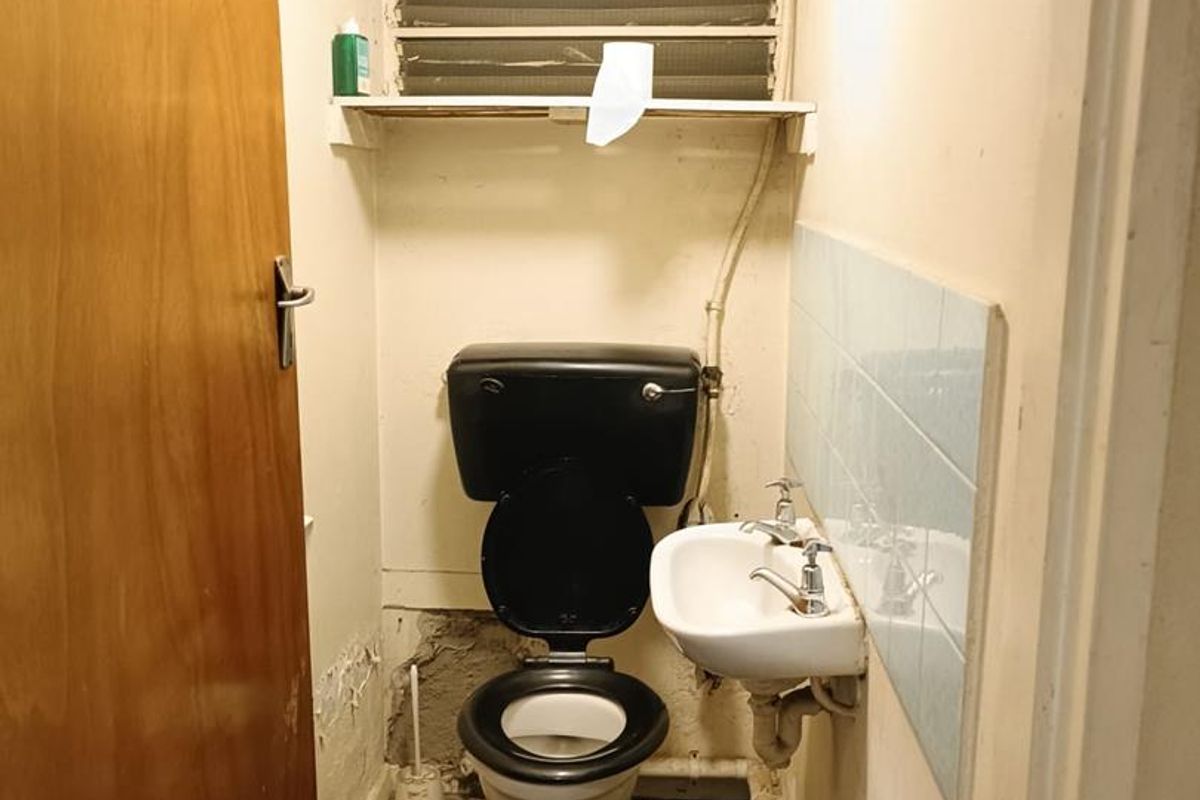 A small, dated bathroom featuring a wall-mounted basin, toilet, and louvered window, requiring significant renovation and repair to the walls and flooring.