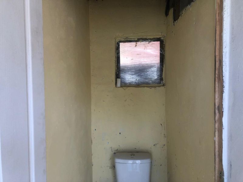 A small, unfinished bathroom space featuring a toilet and a window, requiring significant renovation and cosmetic repairs to the walls and finishes.
