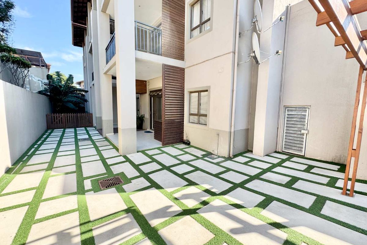A well-maintained rear courtyard featuring a modern paver and artificial turf design, a covered patio area, and timber privacy screening.