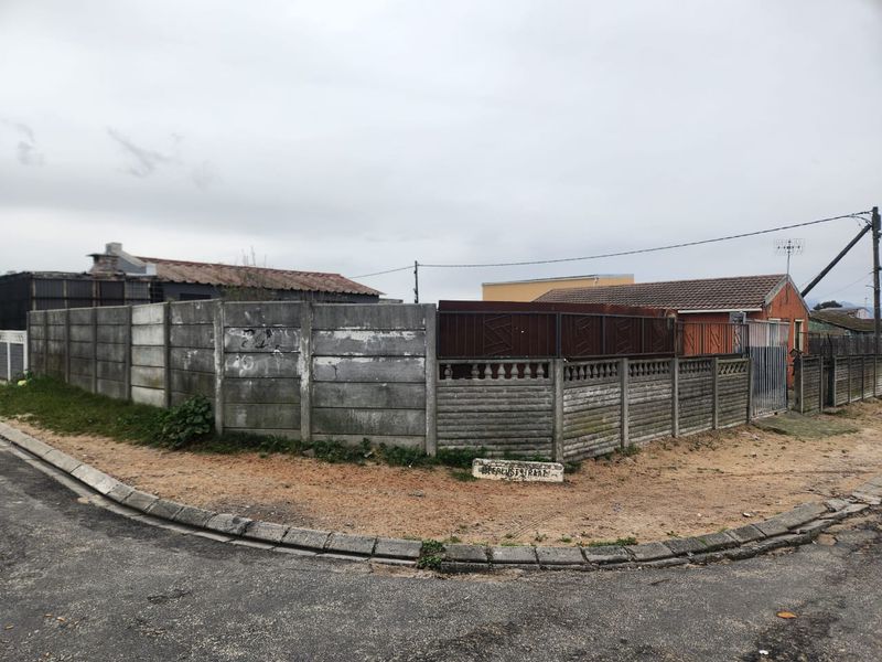 Street view of the property corner featuring a precast concrete boundary wall and metal gate access from the asphalt road.