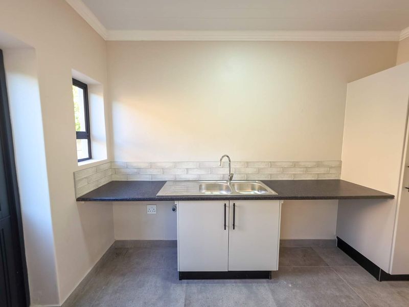 A clean and functional scullery area featuring a double bowl sink, dark laminate countertops, subway tile backsplash, and neutral tiled flooring.