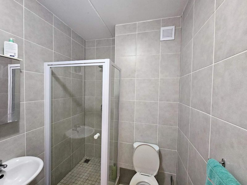 A functional bathroom featuring a glass shower enclosure, pedestal basin, and toilet, finished with neutral wall tiling and integrated ventilation.