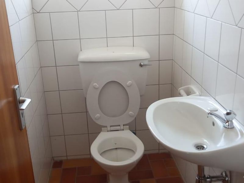 A compact office restroom featuring a standard toilet, wall-mounted basin, and full-height white ceramic wall tiling.