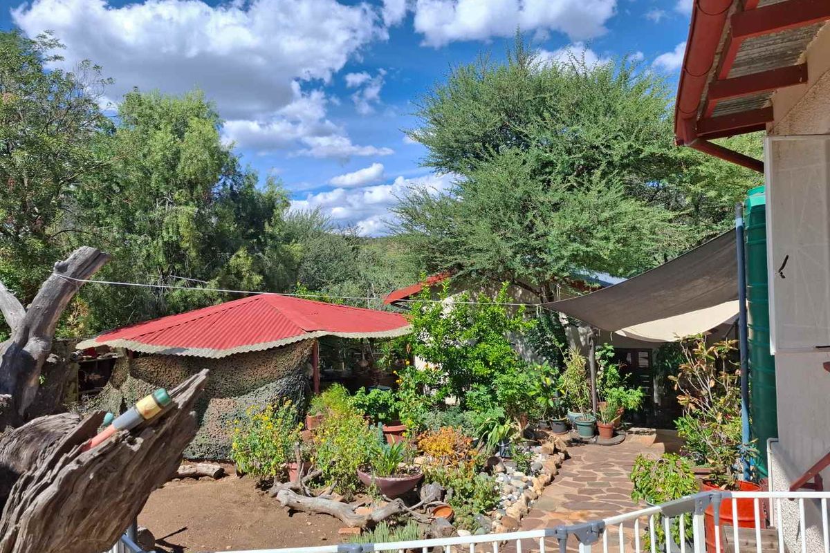 A lush rear garden area featuring a stone pathway, shade sail, and corrugated metal roofing structures, surrounded by mature trees and greenery.