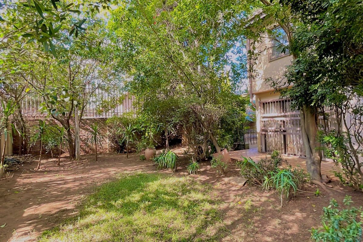 A private rear garden area featuring mature trees, established shrubs, and a paved patio section adjacent to the townhouse structure.