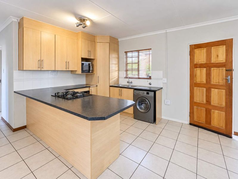 A clean, functional kitchen featuring light wood cabinetry, dark laminate countertops, a gas hob, and tiled flooring with direct access to an exterior door.