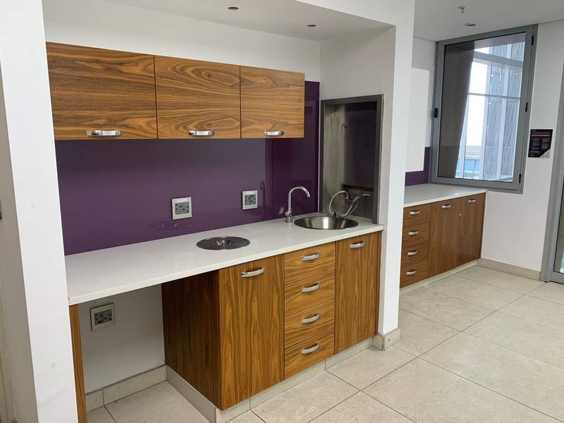 A modern office kitchenette featuring wood-grain cabinetry, stone countertops, a purple glass splashback, and a stainless steel sink.