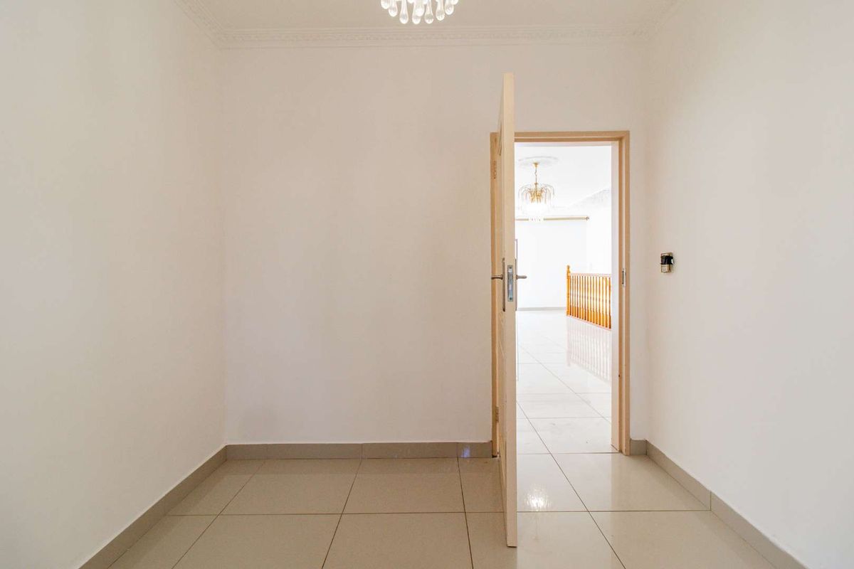 A bright, empty room featuring neutral floor tiles, crown molding, and a central light fixture, with an open doorway leading to an adjacent hallway.