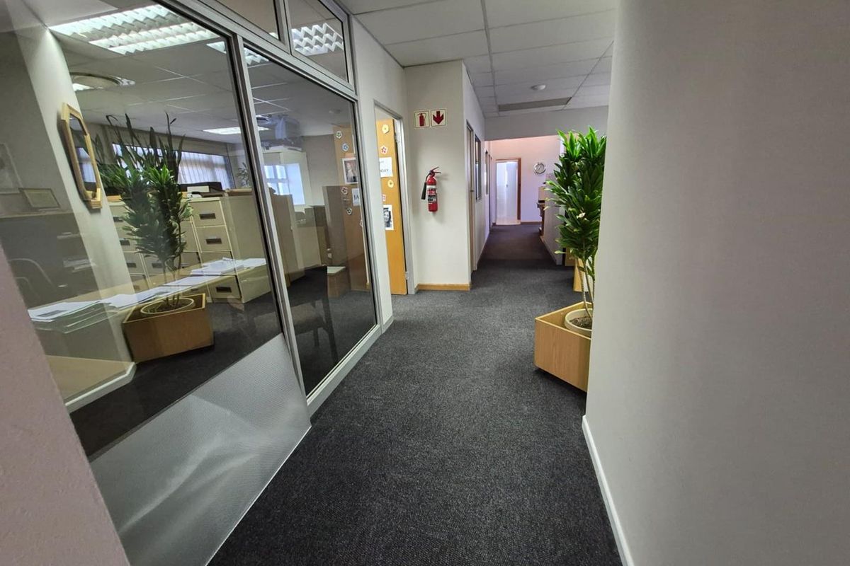 A functional office hallway featuring carpeted flooring, suspended ceilings with integrated lighting, and glass partition walls providing visibility into adjacent office spaces.