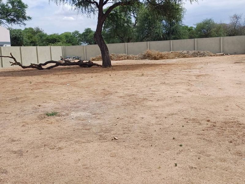 A vacant residential plot featuring a level sandy surface, a mature tree, and secure precast concrete boundary walls.