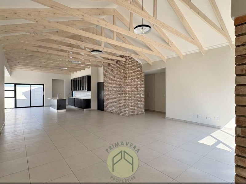 Spacious open-plan living and kitchen area featuring high vaulted ceilings with exposed timber trusses, a rustic brick feature wall, and modern tiled flooring.