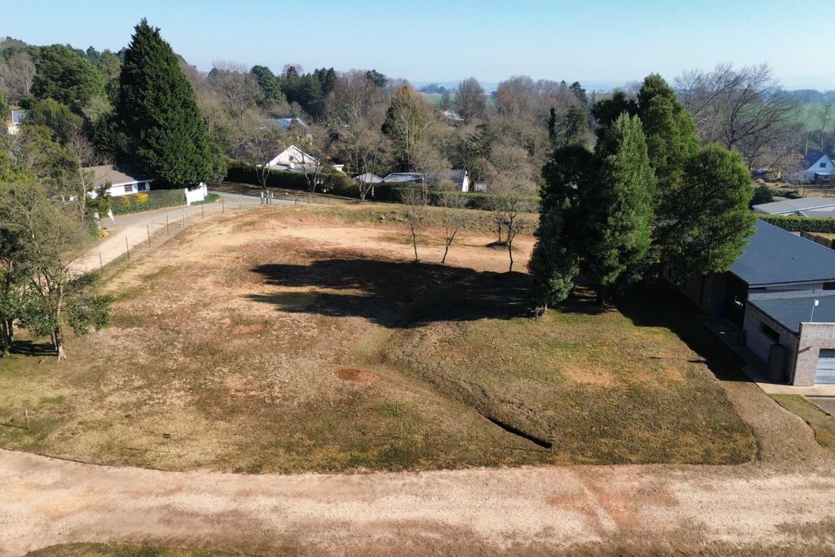 An aerial view of a vacant residential plot featuring established trees, perimeter fencing, and access to a gravel road in a quiet neighborhood.