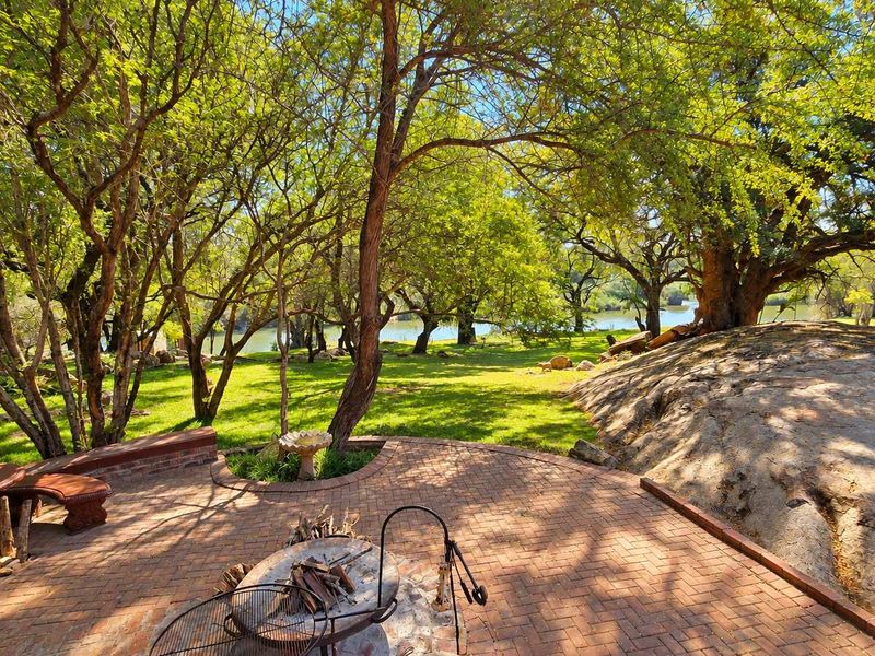 A scenic outdoor patio area featuring brick paving, built-in seating, and a fire pit, overlooking a lush garden with mature trees and a river view.