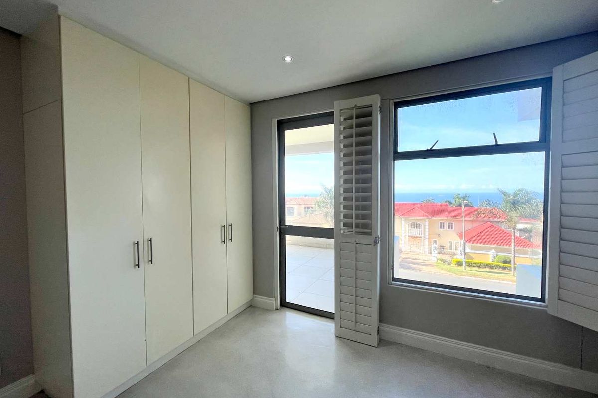 A bright bedroom featuring built-in floor-to-ceiling cabinetry, recessed lighting, and large windows with plantation shutters, offering a view of the surrounding neighborhood and ocean.