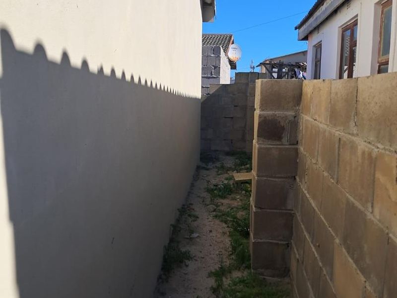 A narrow side passage between the main house structure and an unfinished concrete block boundary wall, featuring unpaved ground with vegetation.