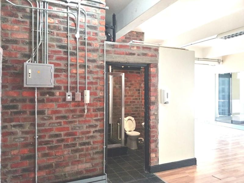 A functional bathroom featuring exposed brick walls, industrial-style electrical conduit, a shower enclosure, and a toilet, set within an open-plan office environment.
