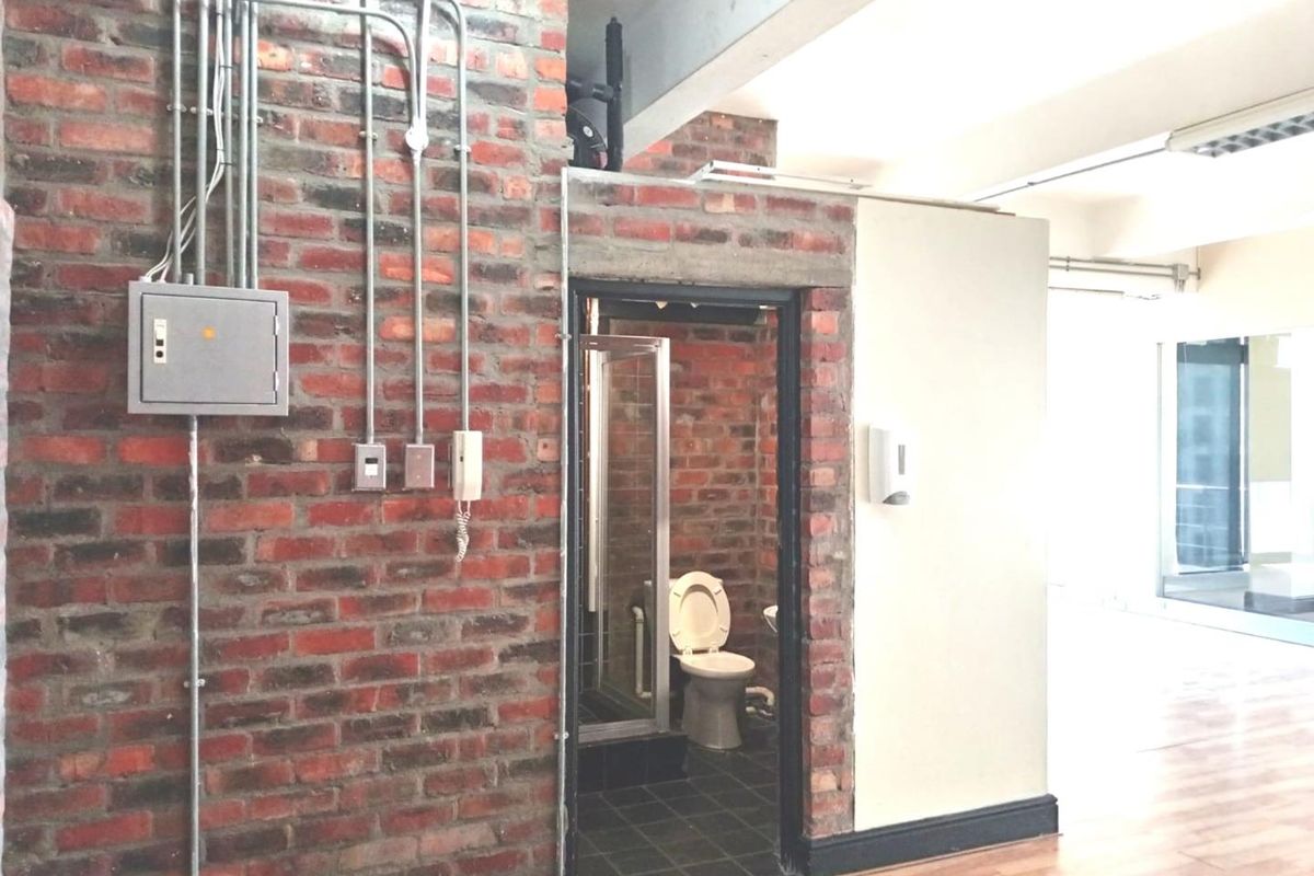 A functional bathroom featuring exposed brick walls, industrial-style electrical conduit, a shower enclosure, and a toilet, set within an open-plan office environment.