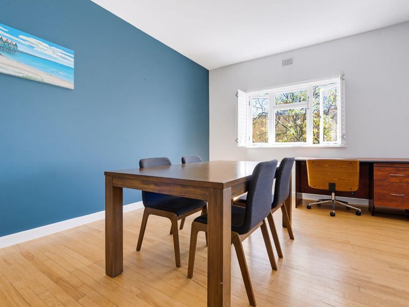 A bright living area featuring hardwood flooring, a blue accent wall, and a large window with fitted shutters providing natural light.