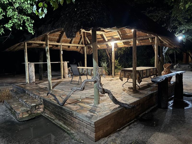 An outdoor thatched-roof entertainment area featuring stone paving, timber support posts, and a built-in bar counter, illuminated for evening use.