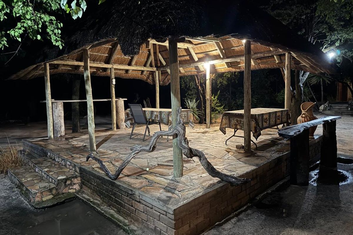 An outdoor thatched-roof entertainment area featuring stone paving, timber support posts, and a built-in bar counter, illuminated for evening use.