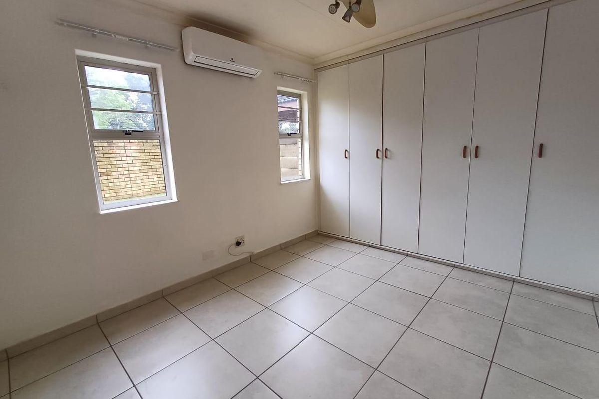 A bright, empty bedroom featuring neutral tiled flooring, built-in floor-to-ceiling cupboards, and a wall-mounted air conditioning unit.