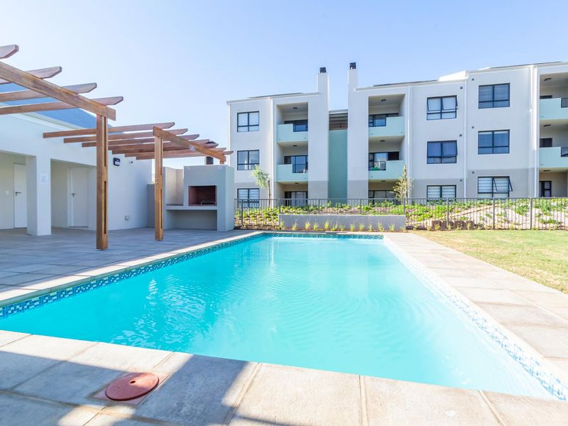 Communal pool area featuring a paved deck, timber pergola, and built-in braai facility, set within a well-maintained apartment complex.
