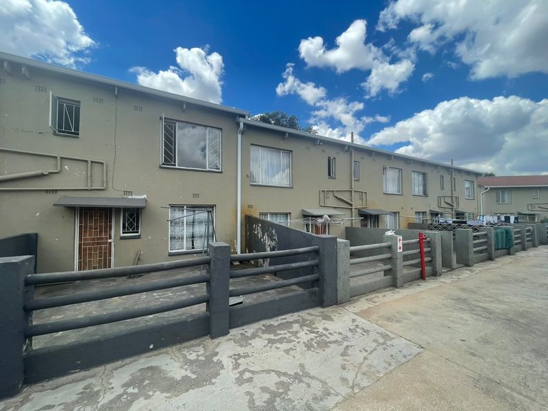 Rear exterior view of a townhouse complex featuring paved courtyards, security gates, and security bars on windows with visible signs of exterior wear.