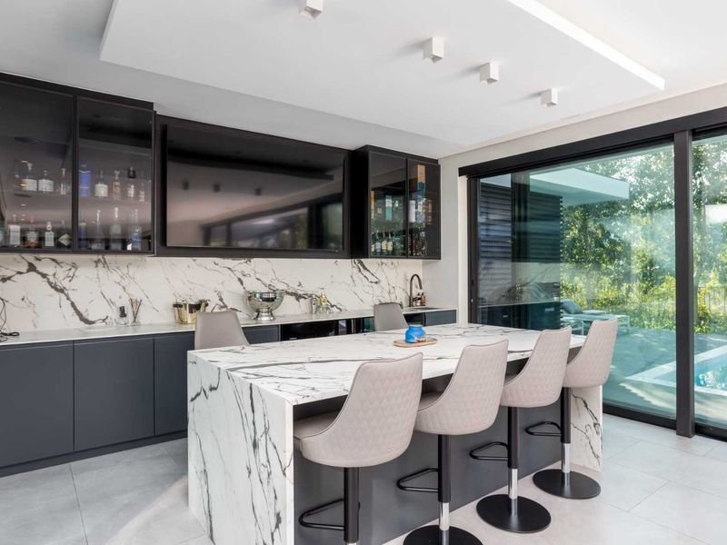 Modern luxury bar area featuring a marble waterfall island, marble backsplash, custom cabinetry with glass display units, and large sliding doors overlooking the pool.