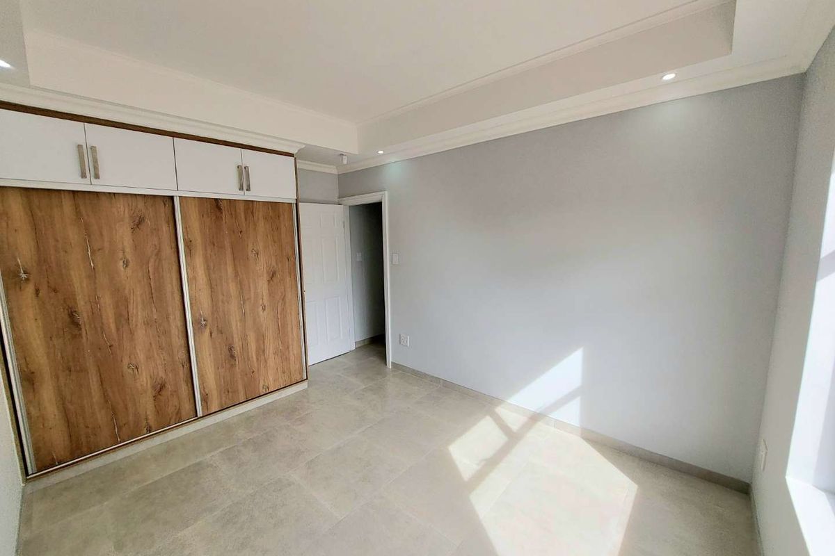A bright, modern bedroom featuring neutral tiled flooring, recessed ceiling lighting, and built-in wooden cabinetry with overhead storage compartments.