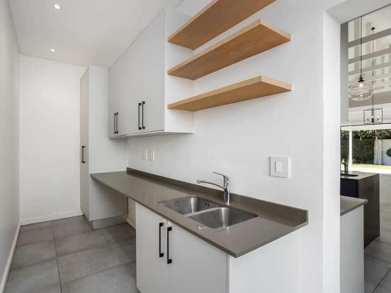 Modern scullery featuring sleek white cabinetry, stone benchtops, double stainless steel sink, and floating timber shelving, providing a clean and functional prep area.