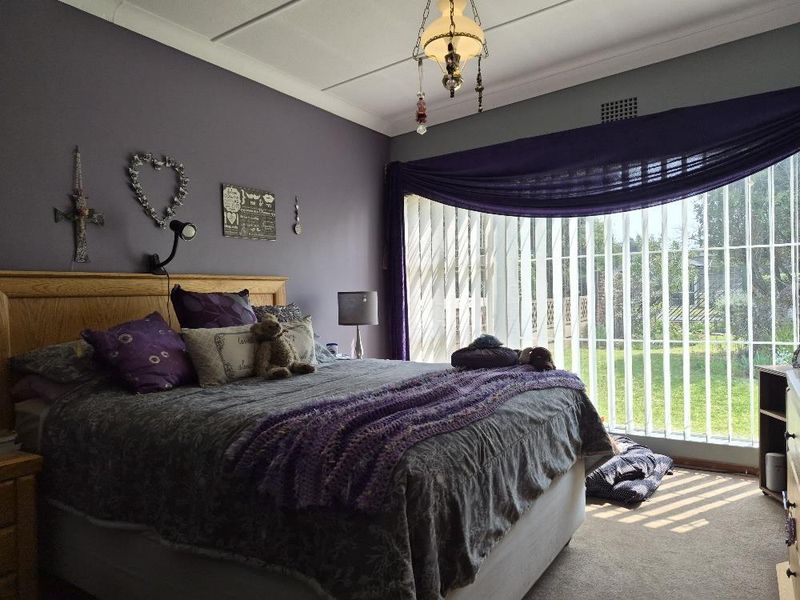 A bedroom featuring carpet flooring, vertical blinds, and a central ceiling light fixture, with a large window providing natural light.