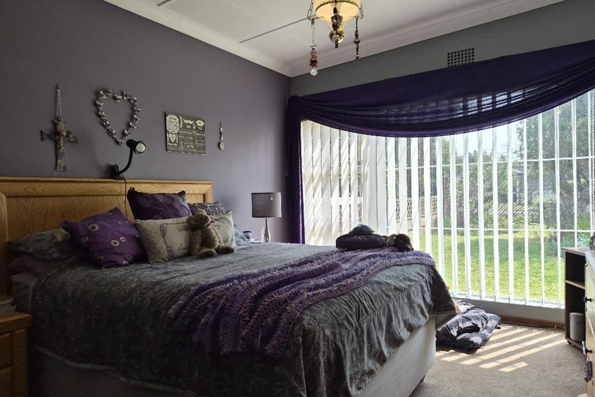 A bedroom featuring carpet flooring, vertical blinds, and a central ceiling light fixture, with a large window providing natural light.
