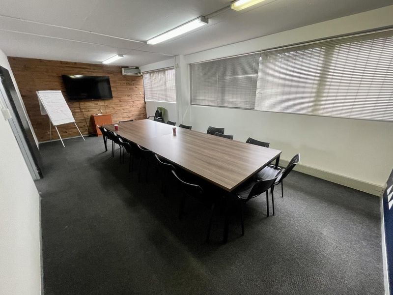 A functional office or boardroom space featuring carpeted flooring, wood-paneled accent wall, large windows with venetian blinds, and overhead fluorescent lighting.