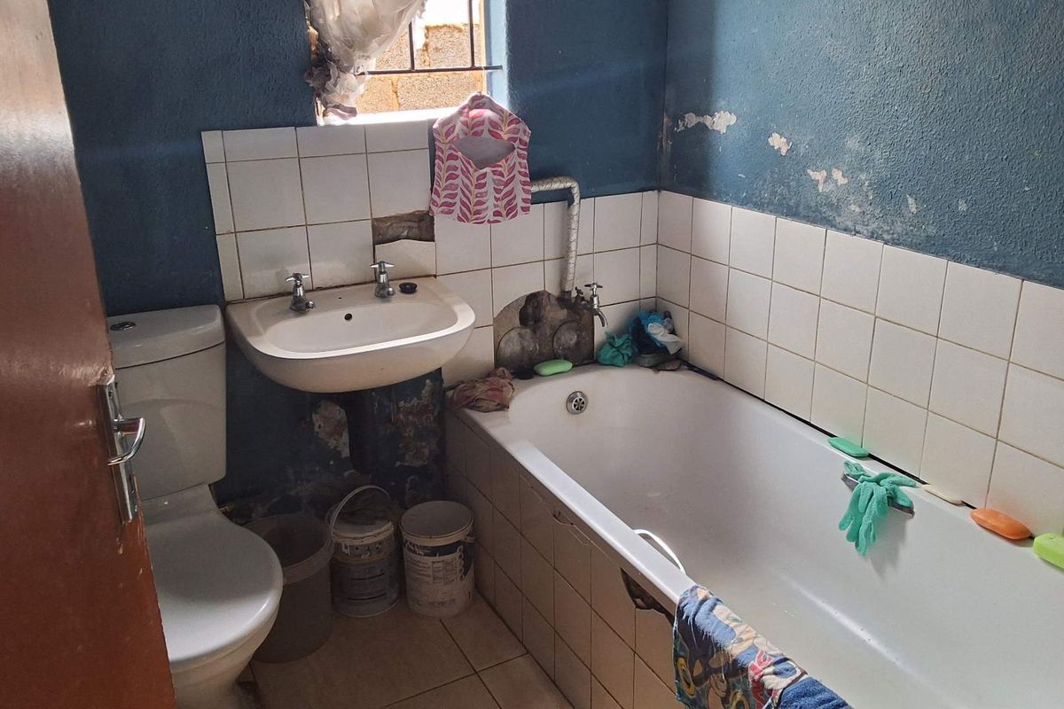 A compact bathroom featuring a standard bathtub, wall-mounted basin, and toilet, currently requiring significant maintenance and cosmetic updates to the wall finishes and tiling.