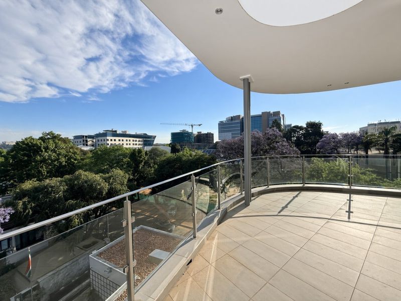 A spacious, modern curved balcony featuring a sleek glass balustrade, tiled flooring, and an expansive urban view.