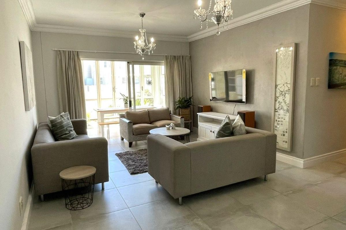 A well-maintained living room featuring large format floor tiles, elegant crown molding, dual crystal chandeliers, and a sliding glass door leading to an outdoor area.