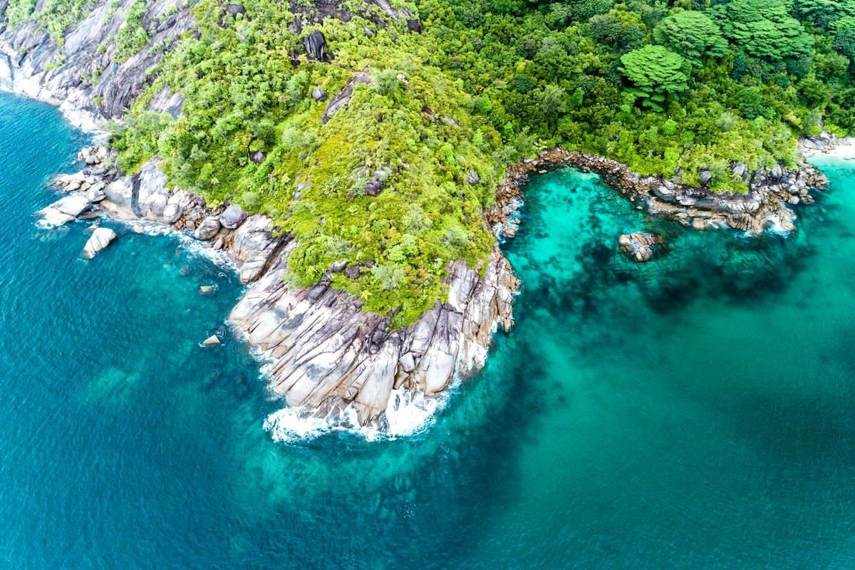 An aerial view of a pristine coastal landscape featuring rocky shorelines, lush tropical vegetation, and clear turquoise ocean waters.