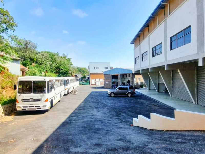 Exterior view of an industrial property featuring a large asphalt yard, multi-story building with roller shutter doors, and a retaining wall.
