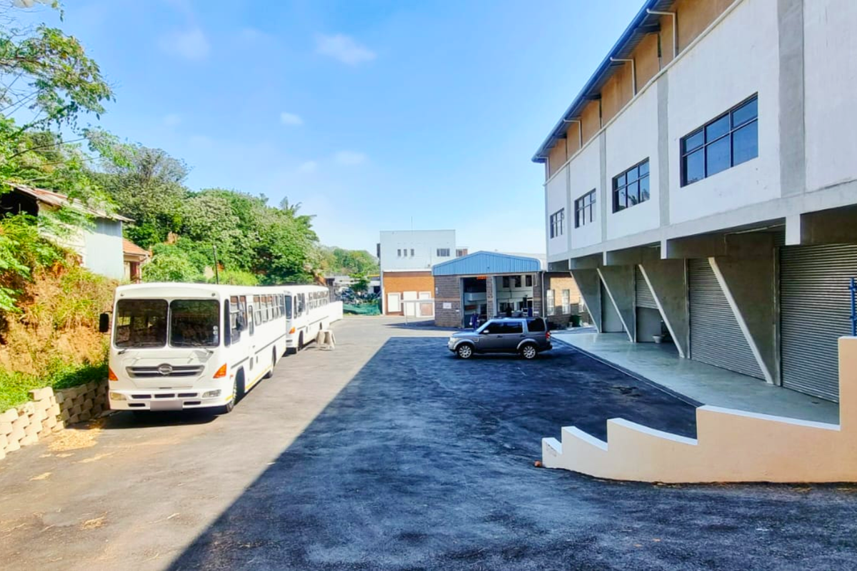Exterior view of an industrial property featuring a large asphalt yard, multi-story building with roller shutter doors, and a retaining wall.