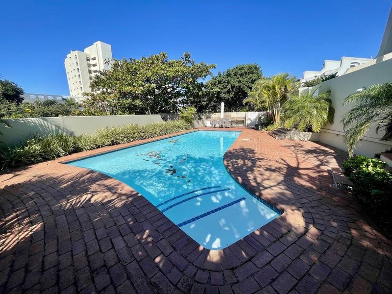 A well-maintained outdoor swimming pool area featuring brick paving, surrounding boundary walls, and established garden landscaping.
