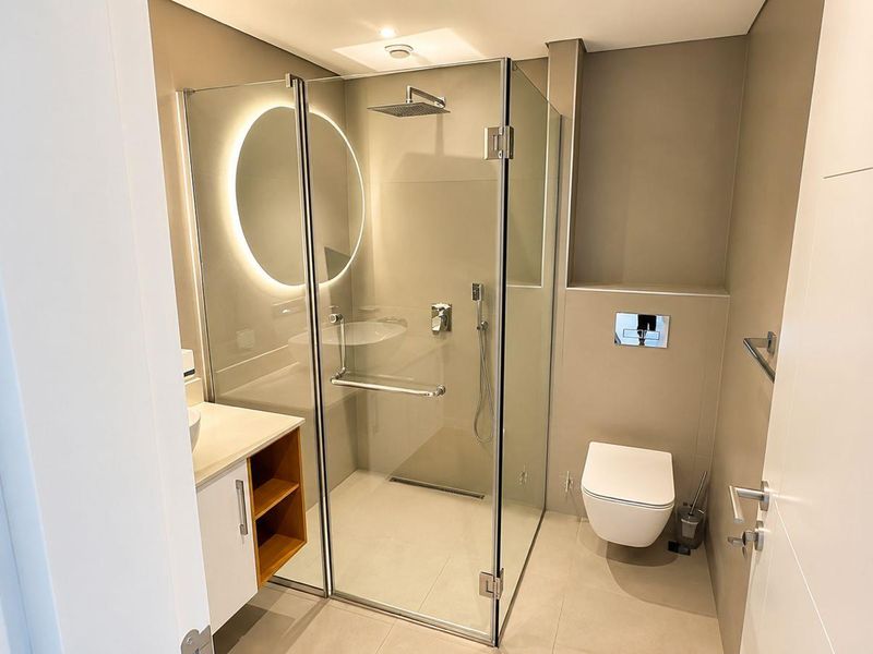 Modern bathroom featuring a glass-enclosed rain shower, wall-mounted toilet, vanity cabinet, and a stylish backlit mirror with neutral tile finishes.
