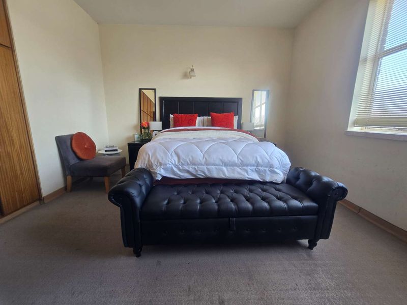 A spacious bedroom featuring a built-in wardrobe, large window with venetian blinds, and neutral wall finishes, offering a functional layout for comfortable living.