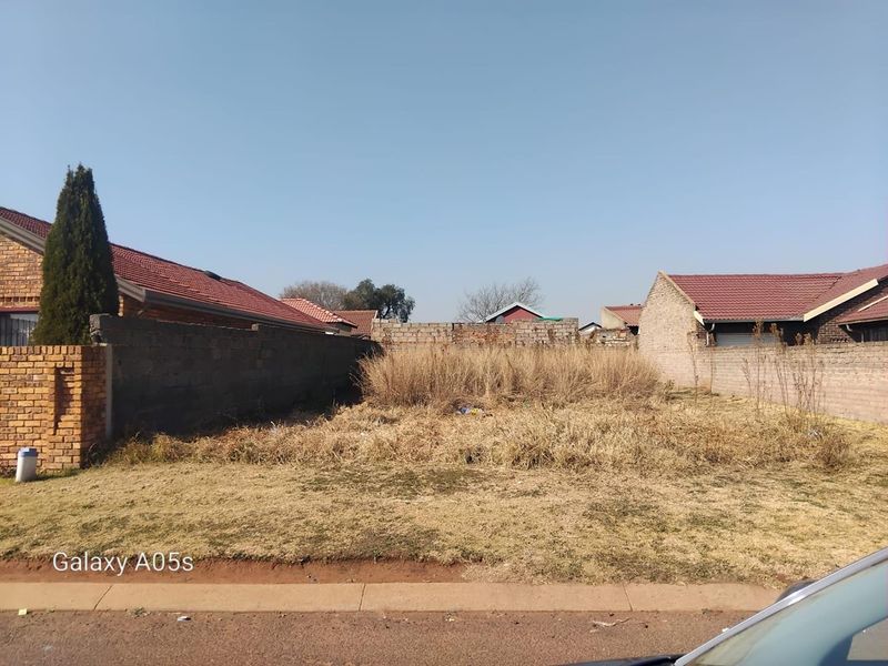 A vacant residential plot enclosed by perimeter brick boundary walls, featuring dry, overgrown vegetation and clear access from the street.