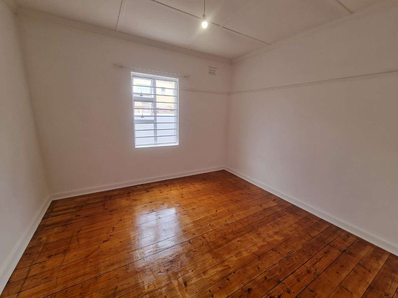 A bright, empty bedroom featuring polished timber flooring, a picture rail, and a steel-framed window with security bars.