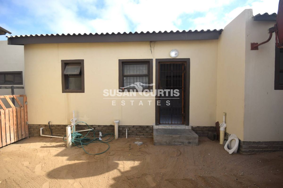 Rear exterior view of the property featuring a simple facade, security gate, and exposed plumbing infrastructure in an unpaved yard area.